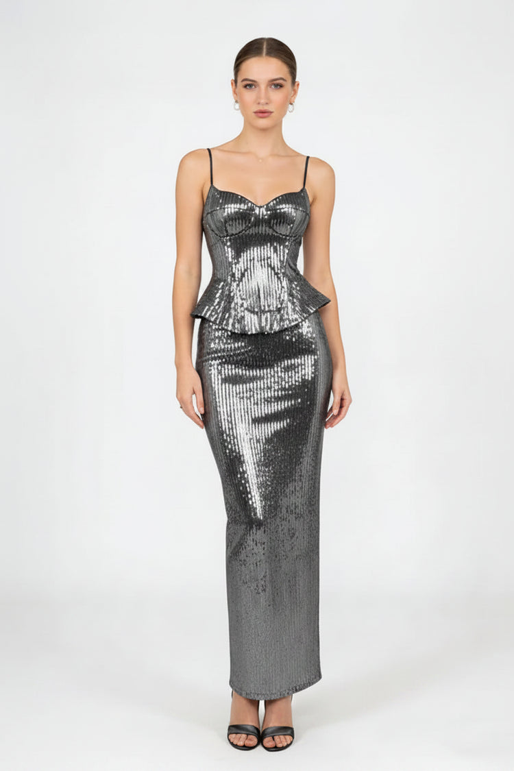 A woman modeling a floor-length, bodycon sequined dress in metallic silver with thin spaghetti straps and a sweetheart neckline, paired with a matching sequined clutch bag and black sandals, against a plain light gray studio background.